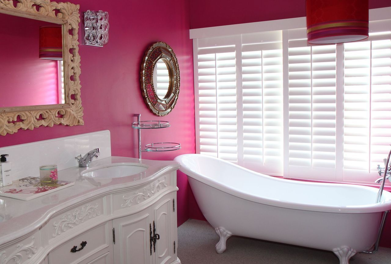 A bathroom with bright pink walls, a white vanity, a pedestal bathtub, and white window shutters.