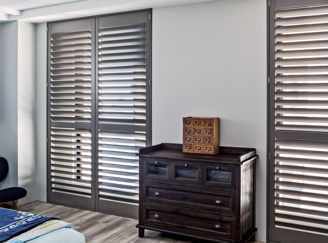 Dark grey plantation shutters cover windows flanking a wooden chest of drawers in a neutral-toned room.
