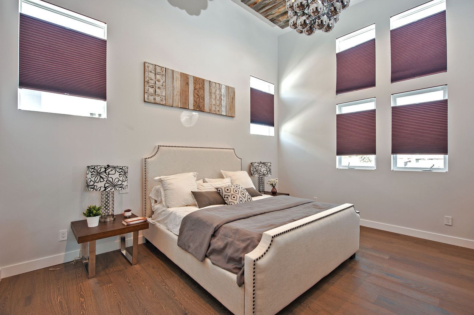 A modern bedroom with a cream upholstered bed, dark wood floor, grey accents, and large windows with plum shades.