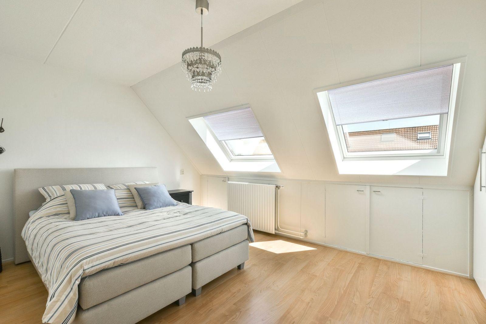A modern attic bedroom featuring a double bed with a striped cover, slanted ceilings, two skylights, and light wood floors.