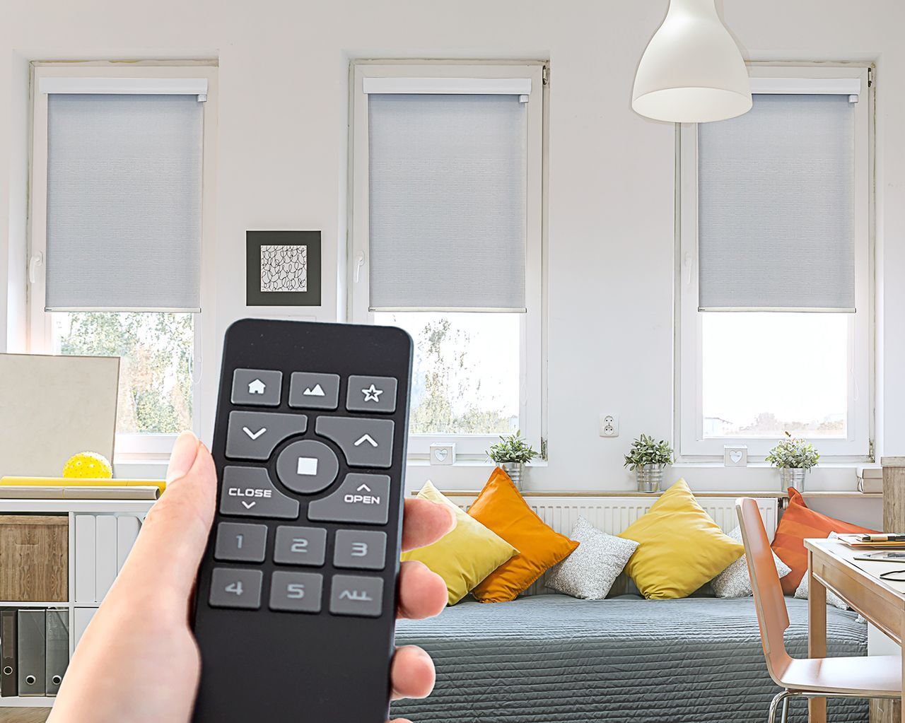 A hand holds a black remote control in a bright room with three windows featuring light gray roller shades.