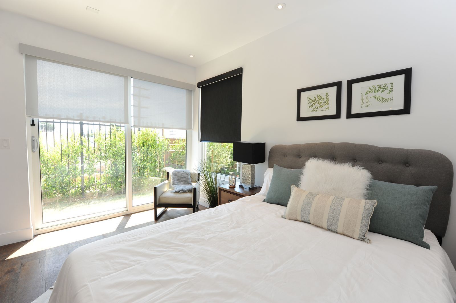 A modern bedroom with a grey tufted headboard, white bedding, two framed prints, and a sliding glass door with a chair.