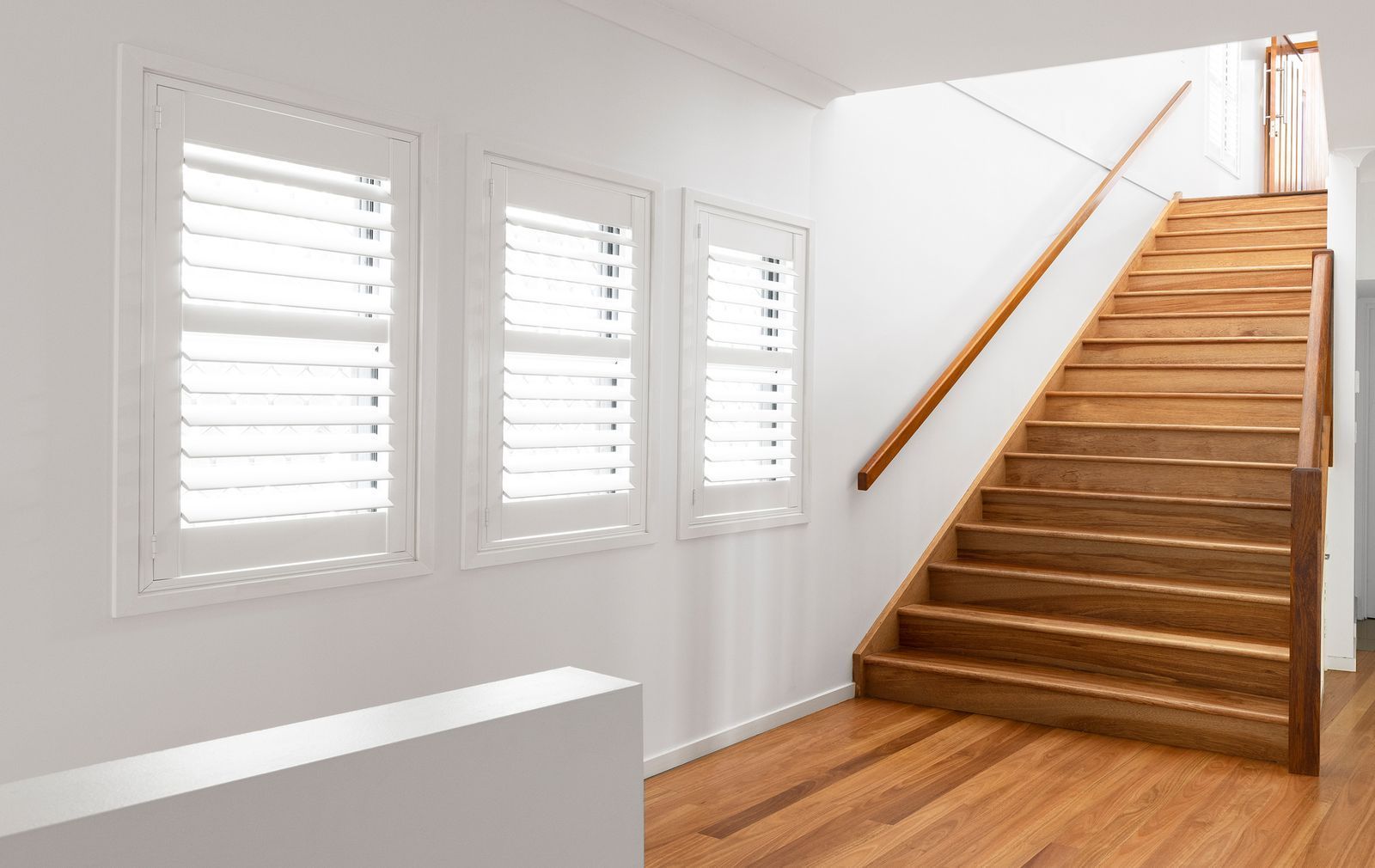 A bright room featuring light wood flooring, white walls with three shuttered windows, and a wooden staircase.