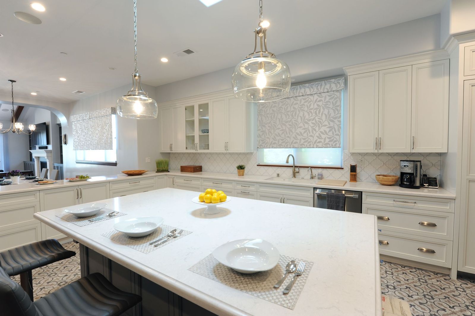 A bright, modern kitchen with white cabinets, a large white island with table settings, and glass pendant lights.