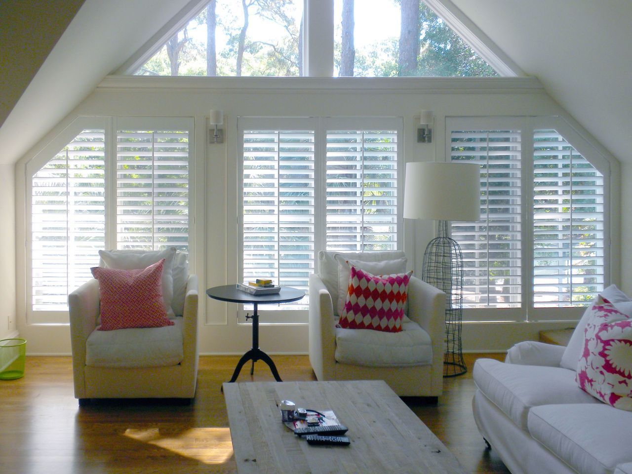 A bright living room with white plantation shutters covering large windows, two armchairs, a couch, and a coffee table.