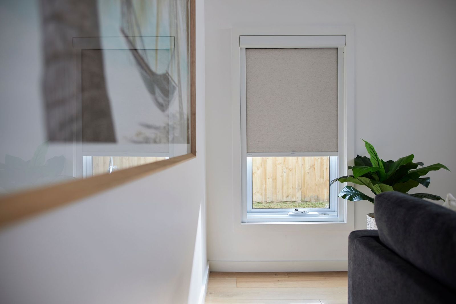 A window with a half-closed beige roller blind, next to a large framed print and a potted plant in a bright room.