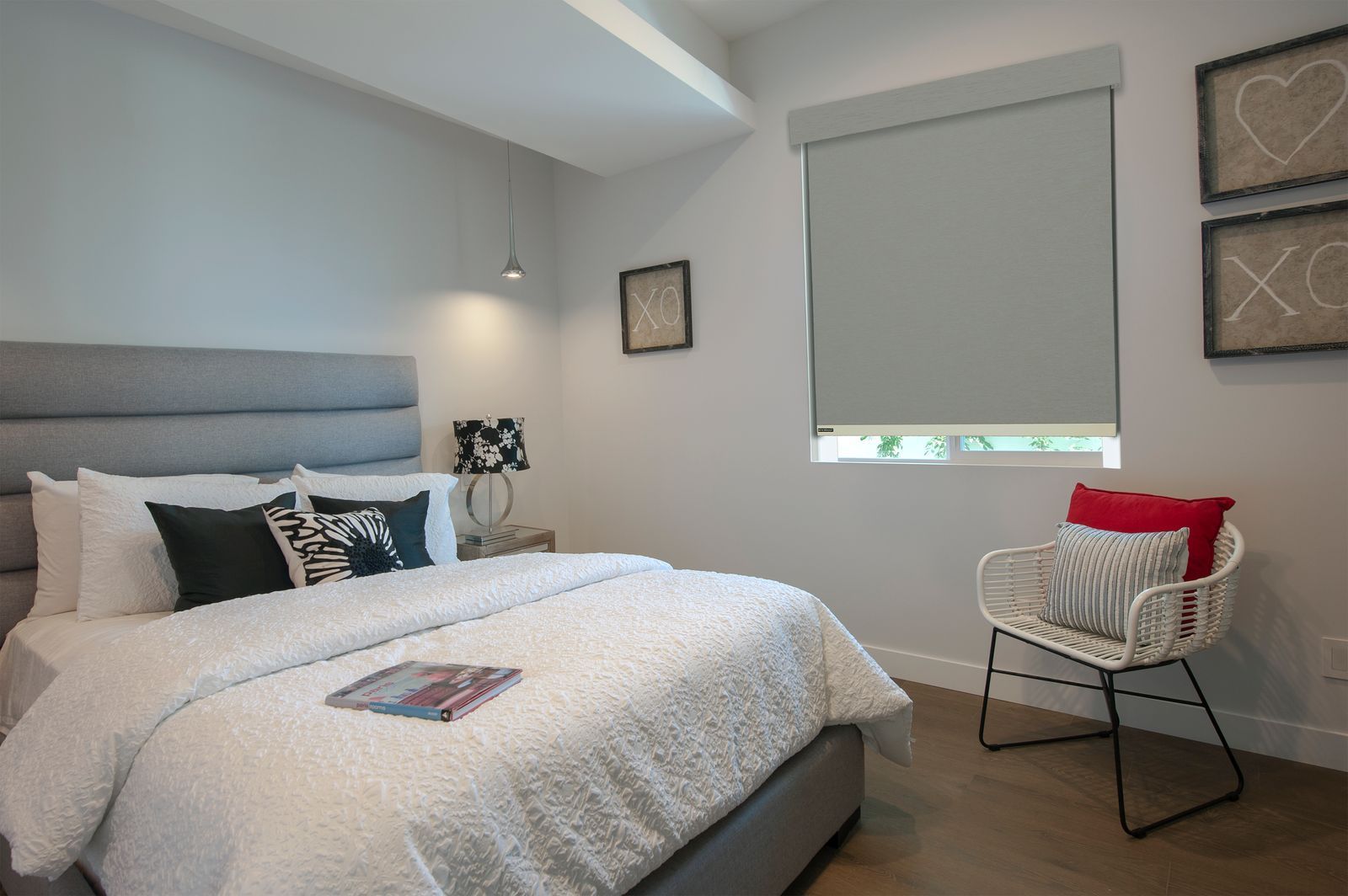 A bright, modern bedroom with a grey upholstered bed, textured bedding, a white wicker chair, and framed wall art.