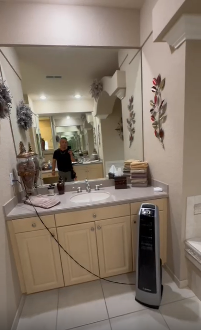 A bathroom vanity with a mirror reflecting a person, a sink, and a portable tower heater on the floor.