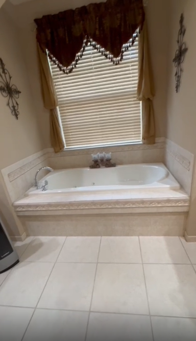 A white jetted bathtub set into a light-tiled deck beneath a window with tan curtains and blinds.