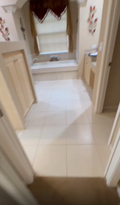 A bathroom with beige tile floors, a white soaking tub under a window with curtains, and light-colored wooden cabinets.