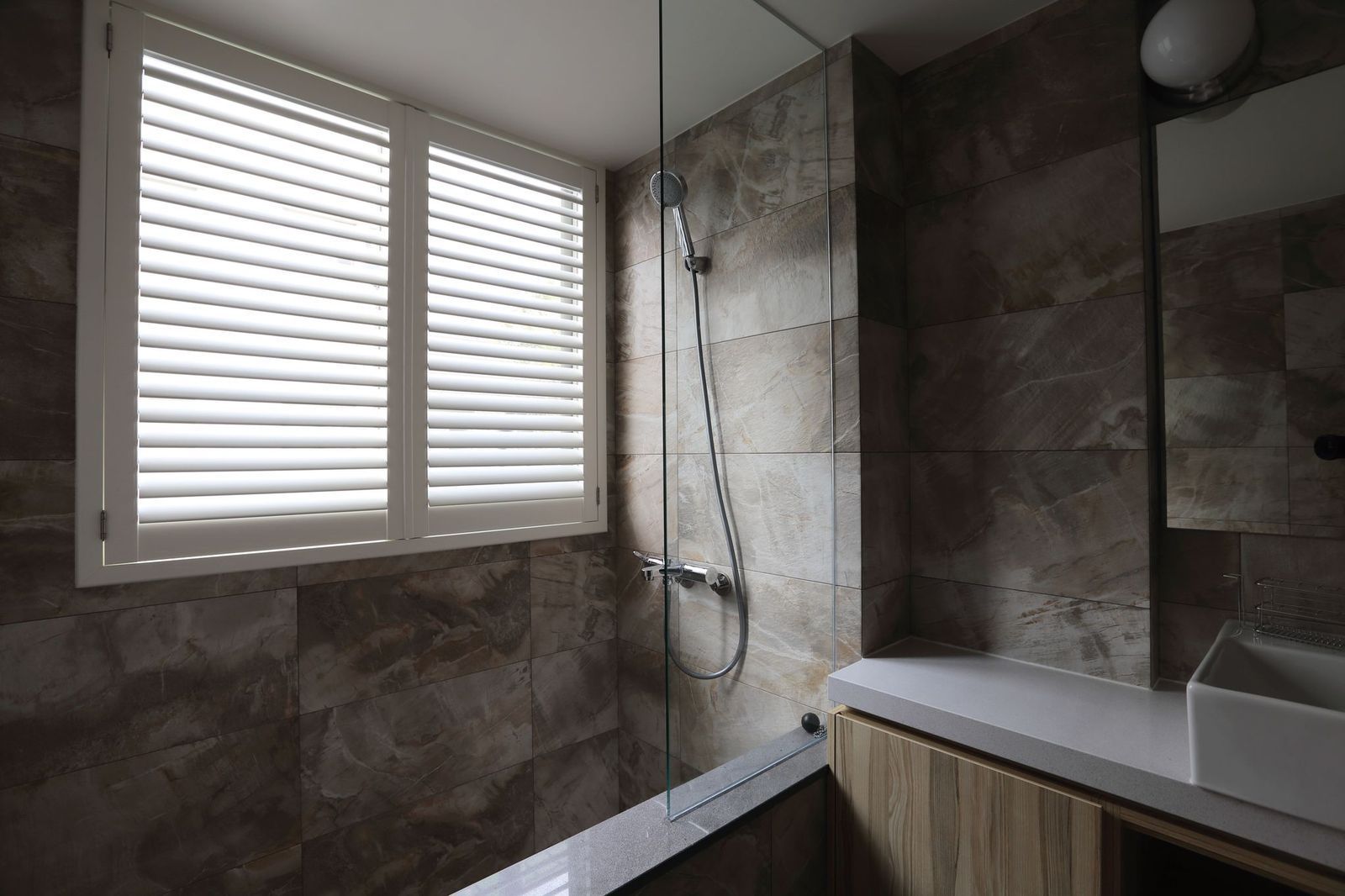 A modern bathroom with dark stone tile walls, a shower behind a glass panel, and a white shuttered window.