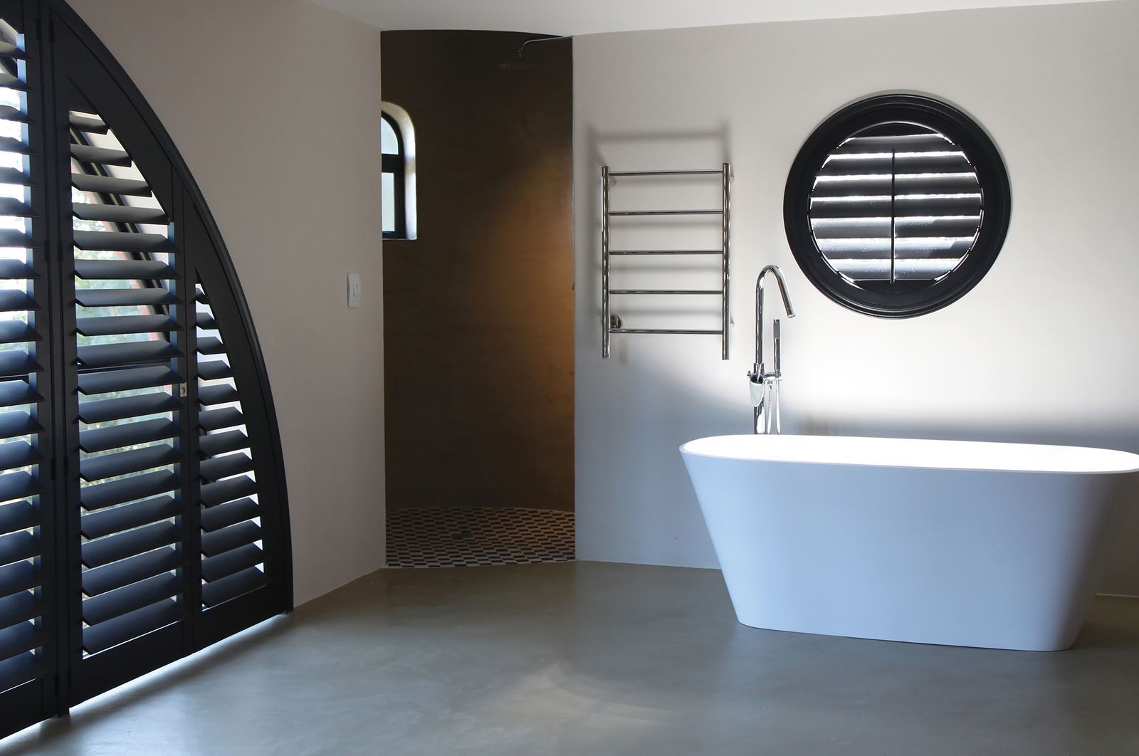 A modern bathroom features a white freestanding bathtub, a silver towel warmer, and black arched and circular shutters.