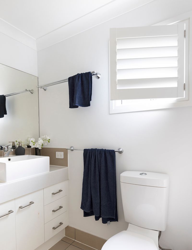 Modern bathroom with white vanity, sink, dark blue towels on racks, white toilet, and a window with white shutters.