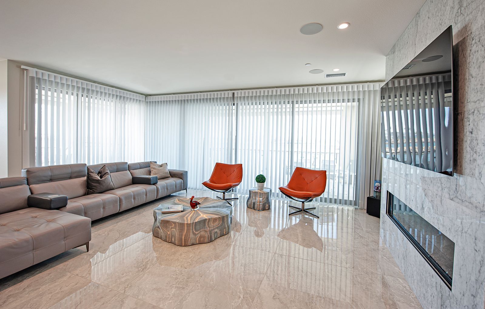 A modern living room with a large gray sectional sofa, two orange armchairs, a stone coffee table, and a wall-mounted TV.