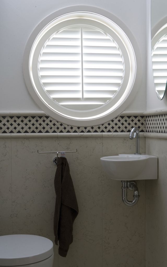 A bathroom with a circular window, white louvered shutters, a wall-mounted sink, and a dark towel hanging on a rack.
