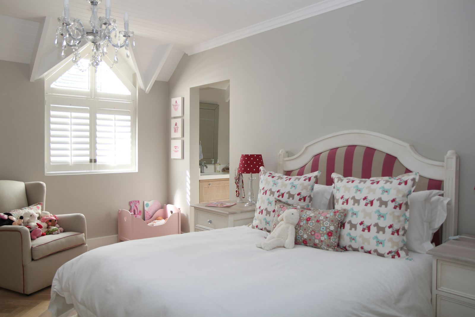 A brightly lit children’s bedroom with a white bed, red-striped headboard, floral pillows, an armchair, and a chandelier.