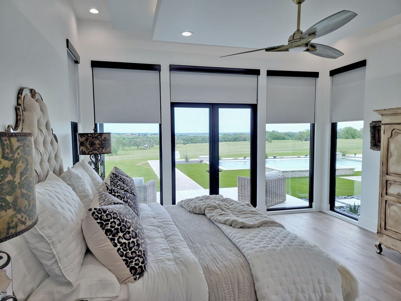 A bedroom with a tufted headboard, patterned pillows, a ceiling fan, and large windows overlooking a pool and landscape.