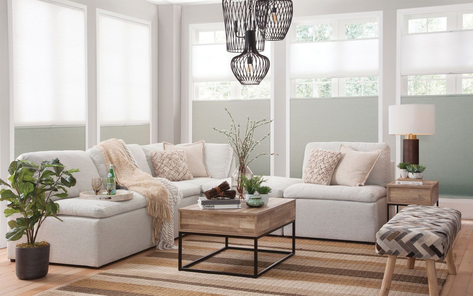 A cozy living room with a white sectional, wood coffee table, patterned bench, and light-filtering shades on the windows.