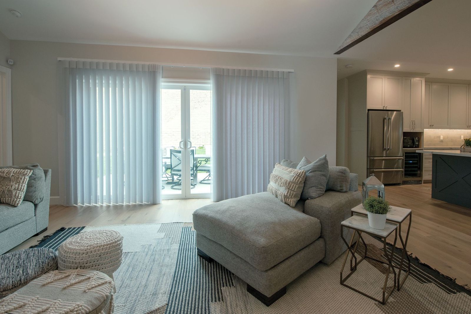 A modern living room with a grey sofa, matching ottoman, and two side tables next to large windows with vertical blinds.