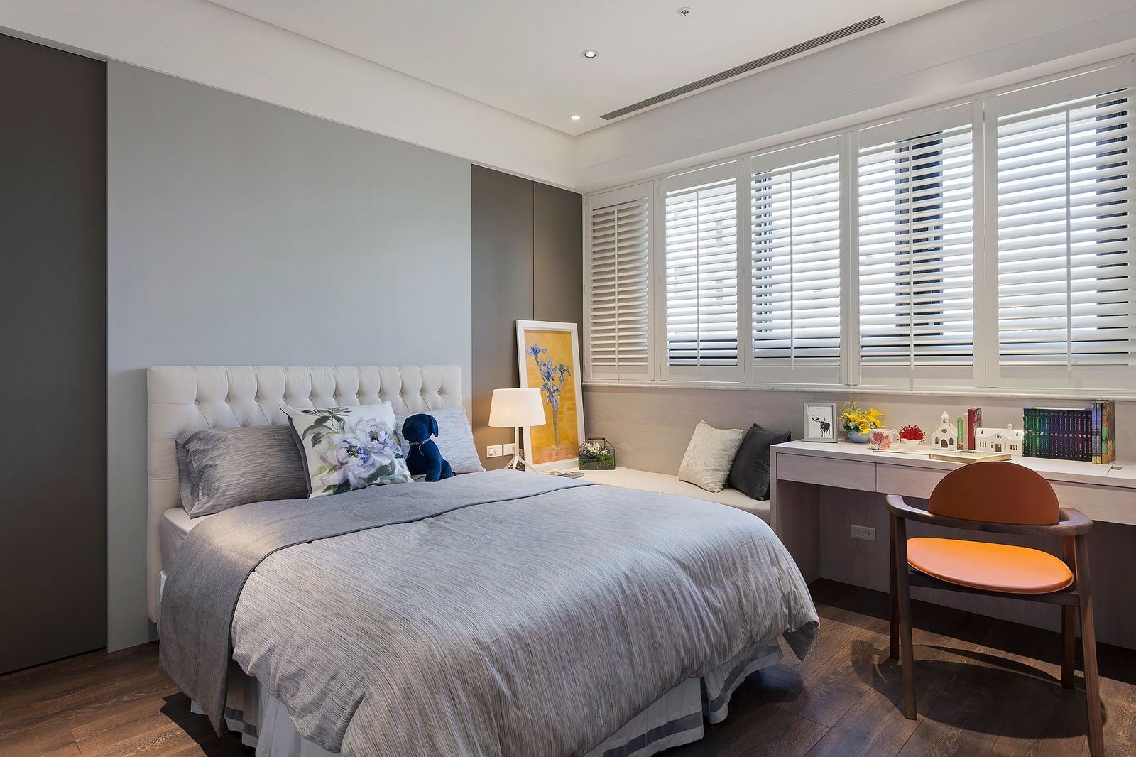 A modern bedroom with a grey tufted bed, a desk area with a chair, and plantation shutters covering large windows.