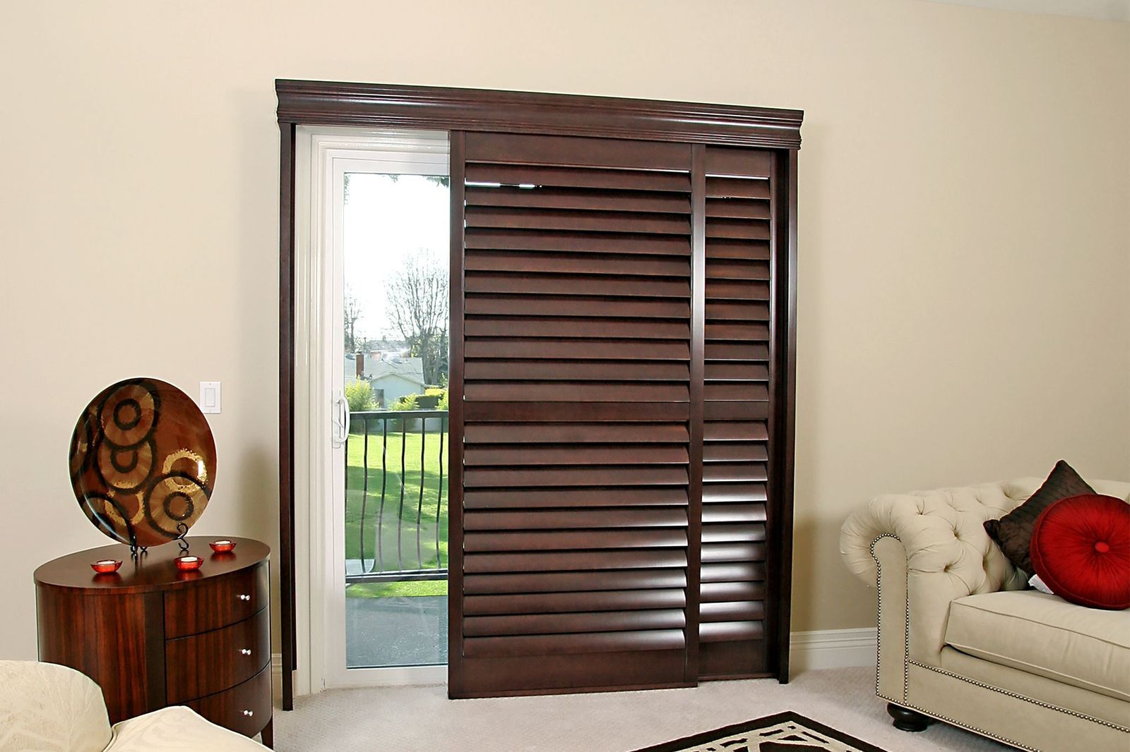 Dark wood sliding shutter panels covering a glass patio door in a beige living room with a couch and side table.