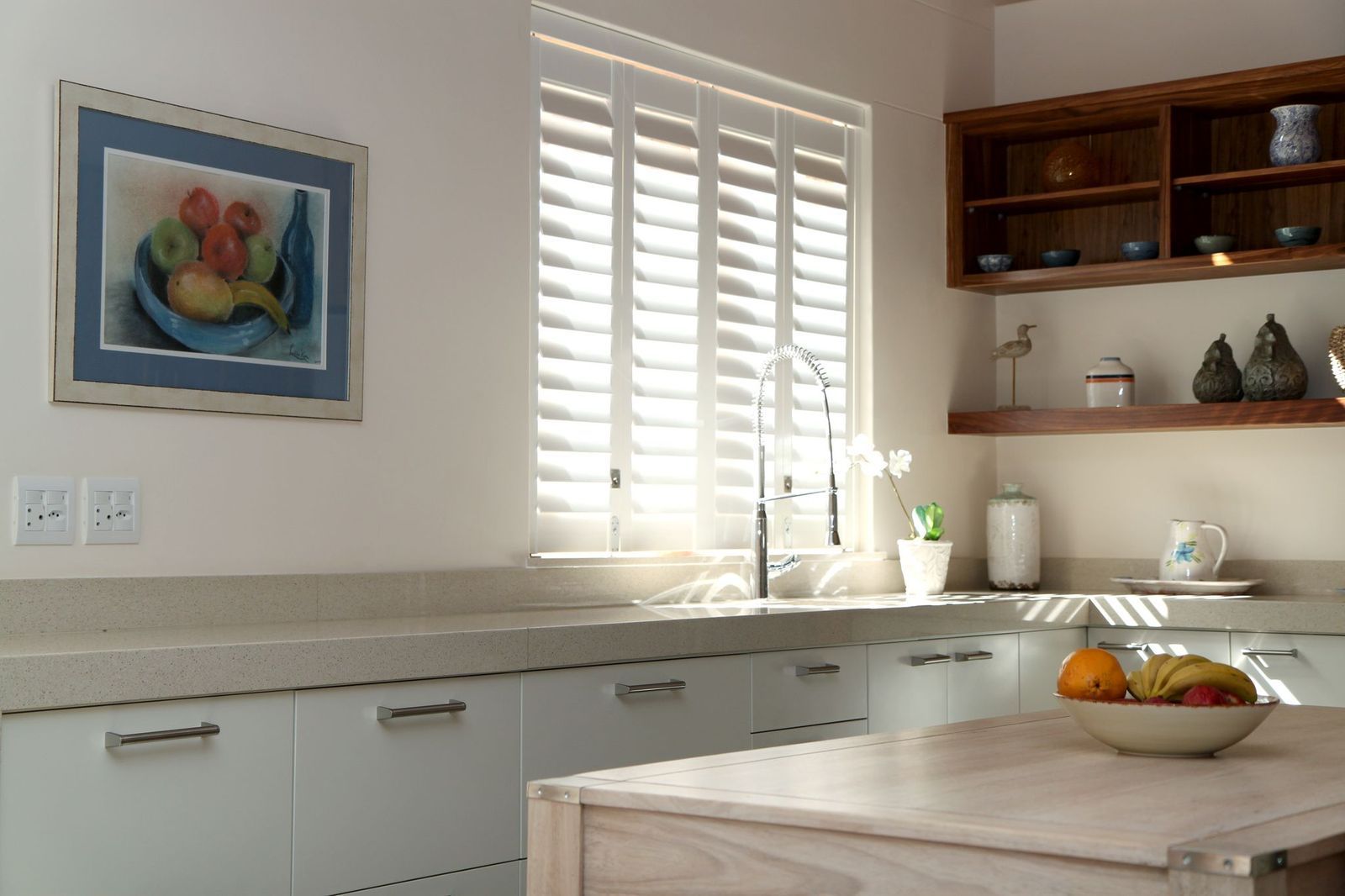 A bright kitchen with white cabinets, a light countertop, wood shelving, a window with white shutters, and a fruit bowl.