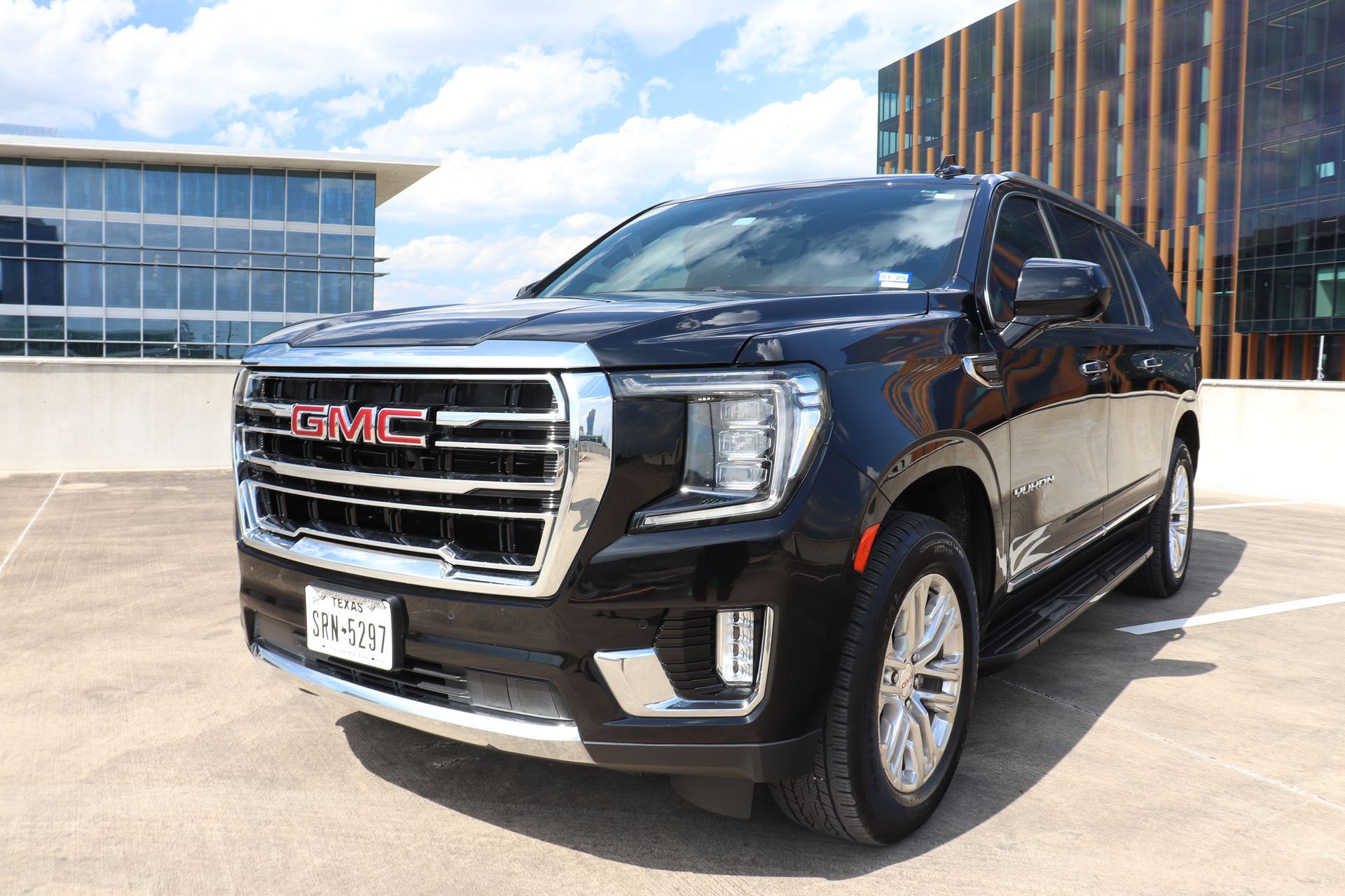 Black GMC Yukon SUV parked on a rooftop, sunny day. Modern buildings in the background.