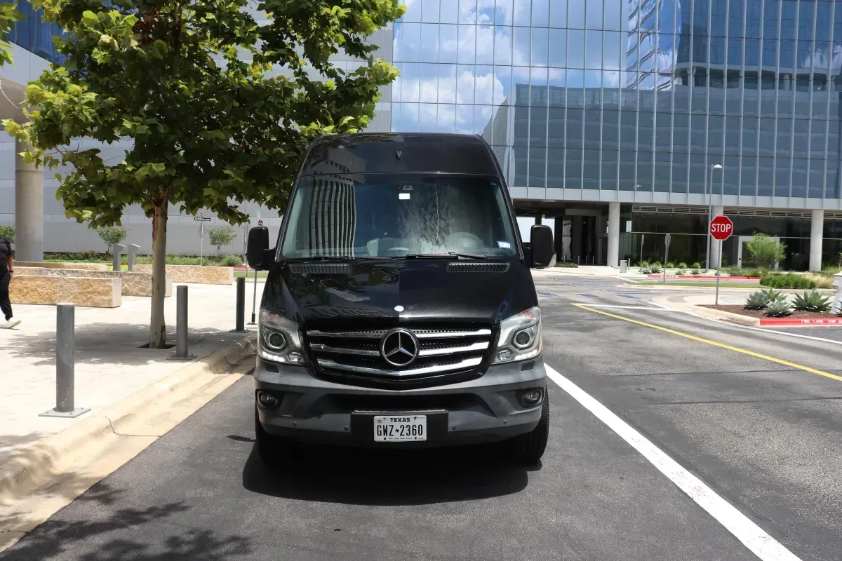 Black Mercedes-Benz Sprinter van parked on a street in front of a modern building with a tree on the left.