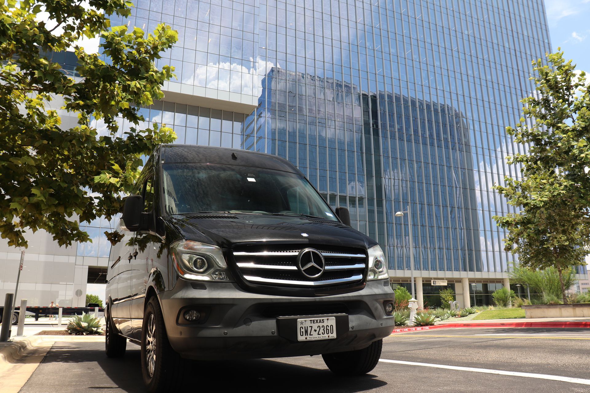 Black Mercedes-Benz van parked on a street in front of a modern glass building; sunny day.