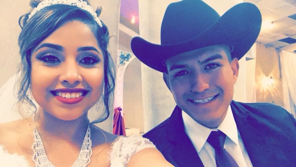 Bride and groom smiling at the camera, bride in a wedding dress and veil, groom in a cowboy hat and suit.