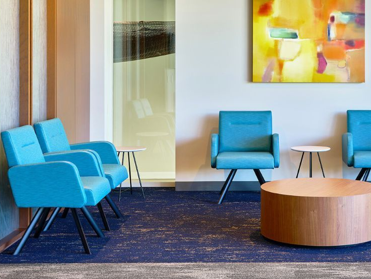 Blue waiting room chairs, small side tables, and round coffee table on blue carpet; painting on the wall.