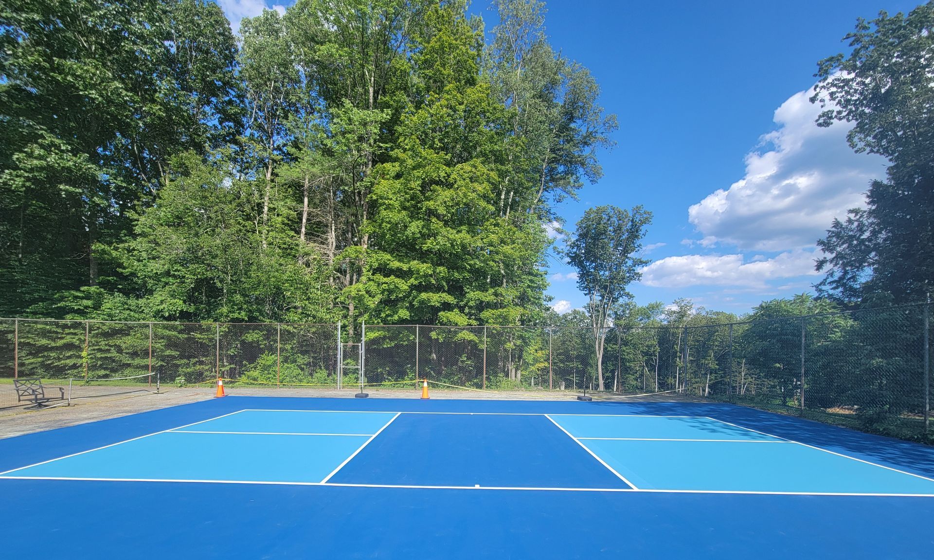Blue outdoor basketball court with a forest backdrop under a blue sky.