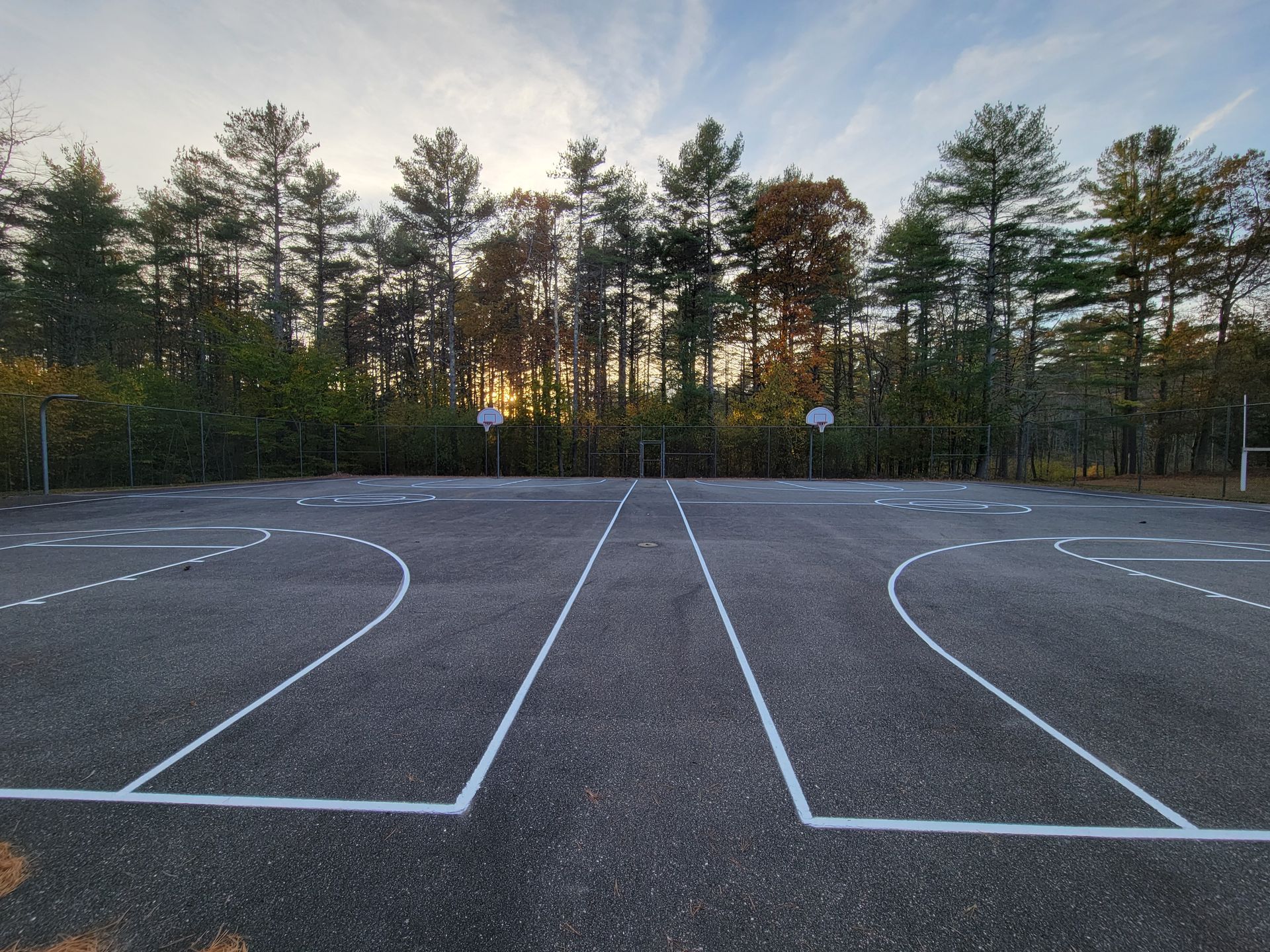 Outdoor basketball court with white lines, hoops, and trees at sunset.