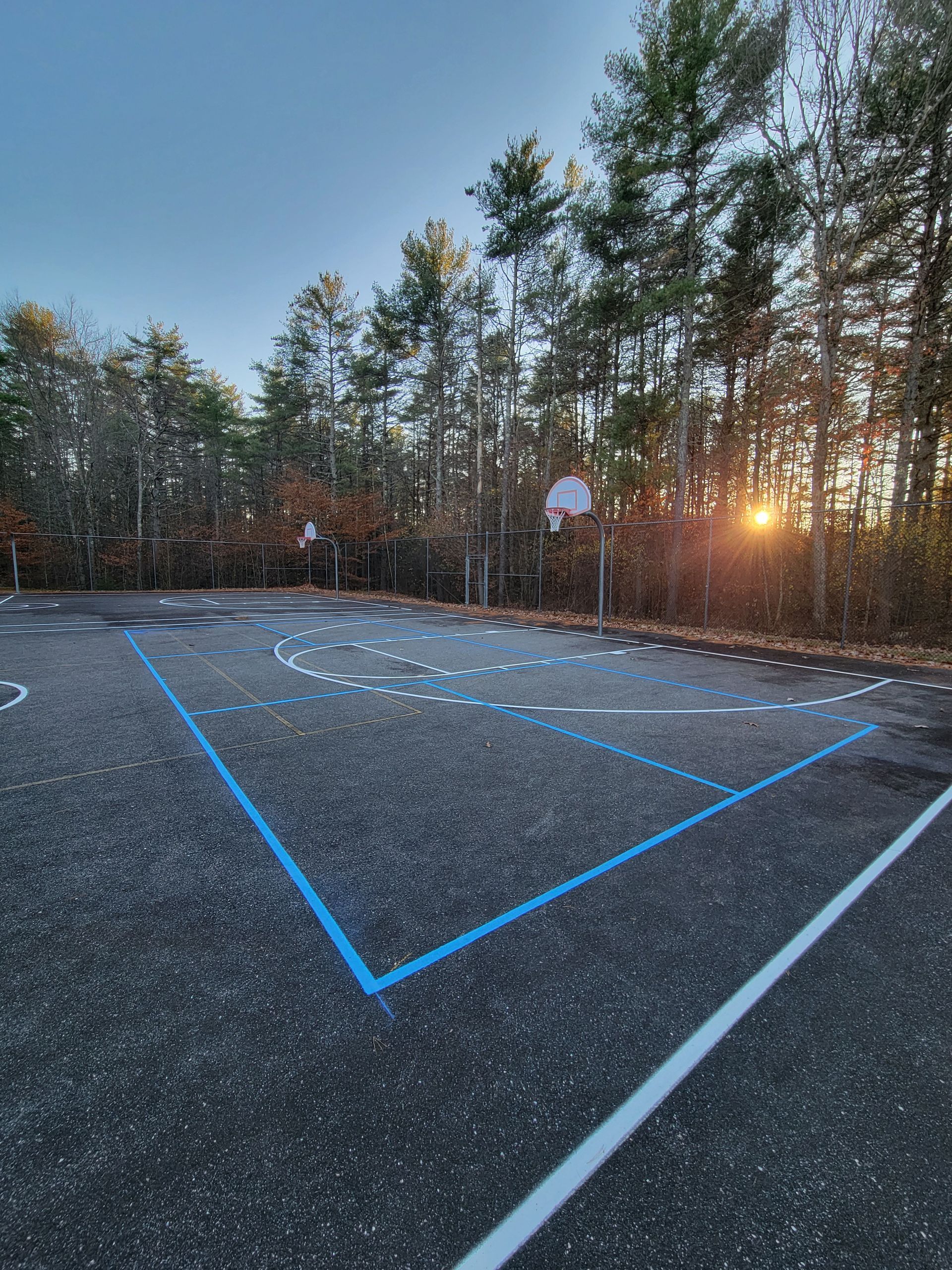 Basketball court with blue and white lines on asphalt, with a backboard and trees in background.