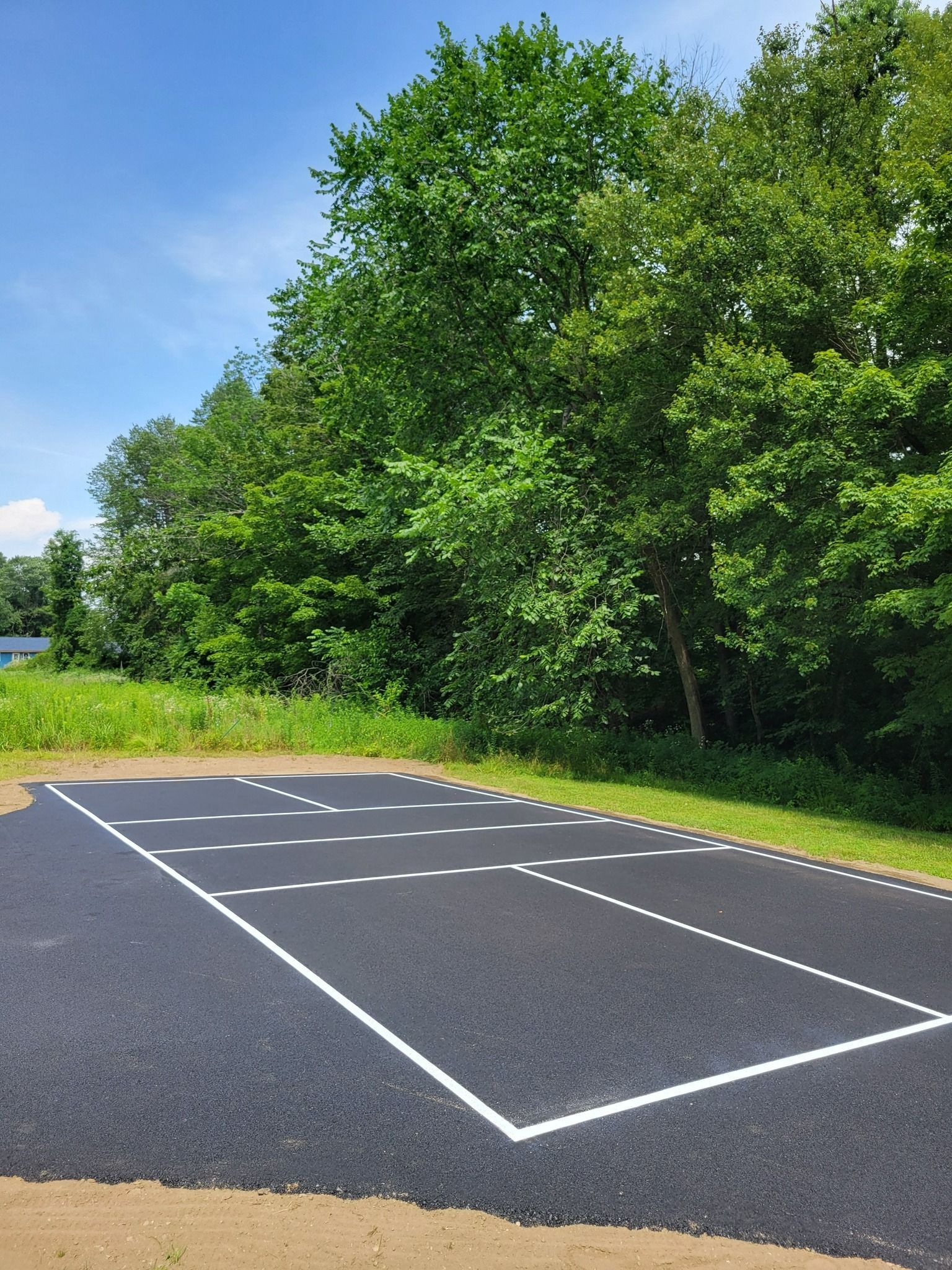 Black asphalt surface with white lines and dots, set in a grassy area near trees.