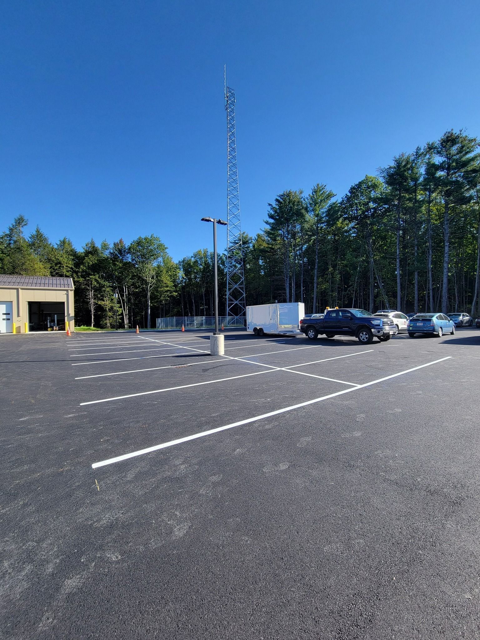 Empty asphalt parking lot with white parking lines. Cars, cell tower, and trees are in the background under a blue sky.