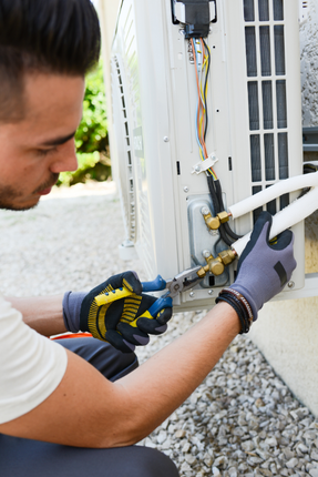 A man wearing gloves is working on an air conditioner outside.