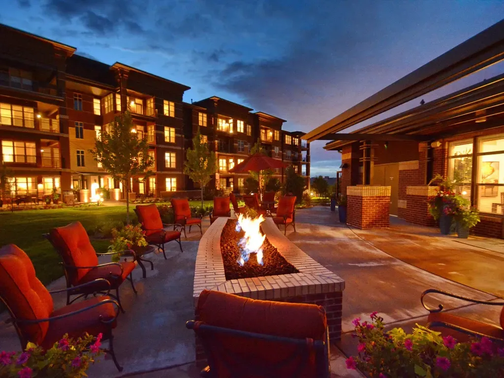 Courtyard fire pit with red chairs outside a lit apartment building at dusk.