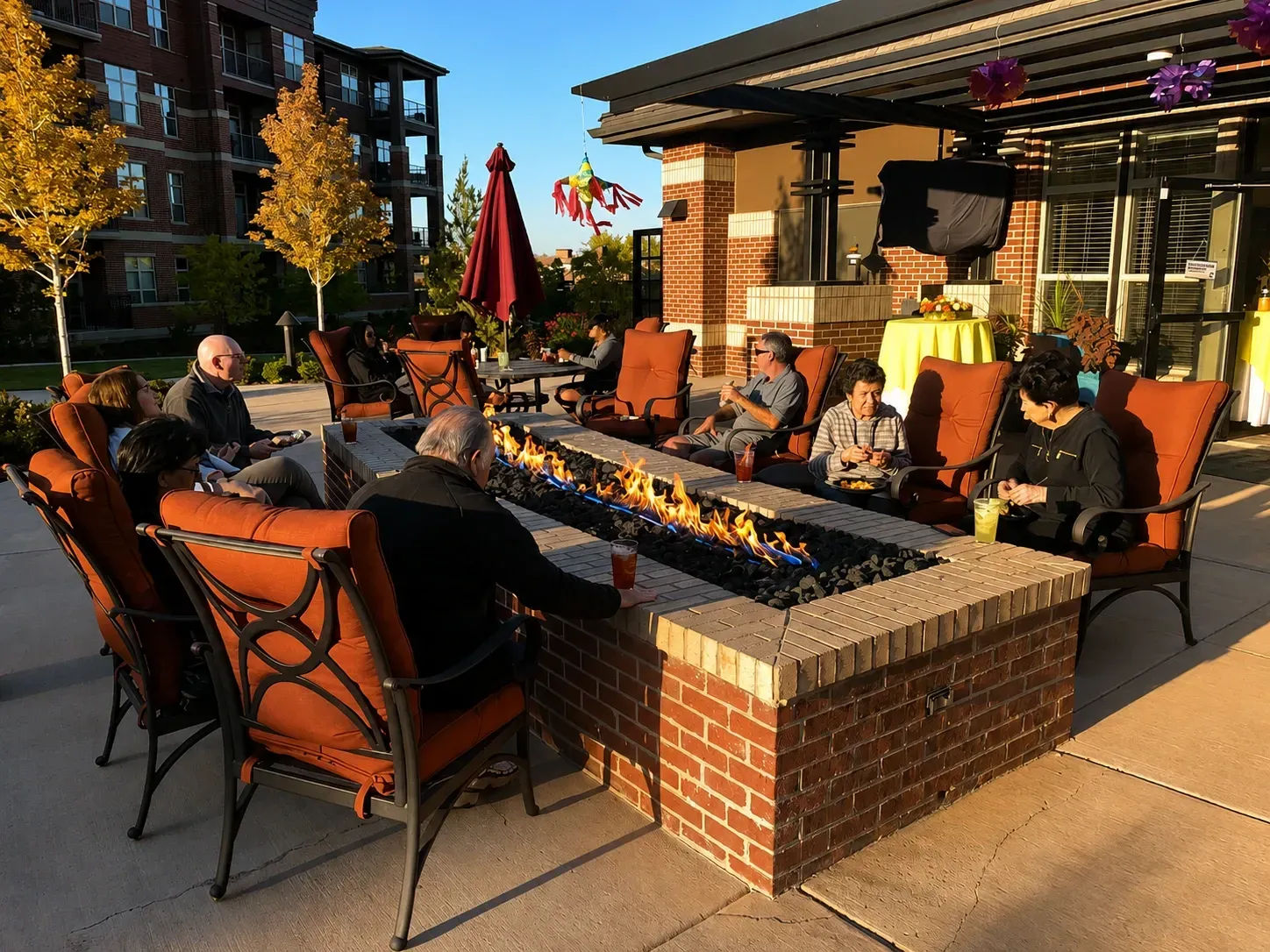 People seated around a brick fire pit on a sunny apartment patio with lounge chairs and umbrellas.