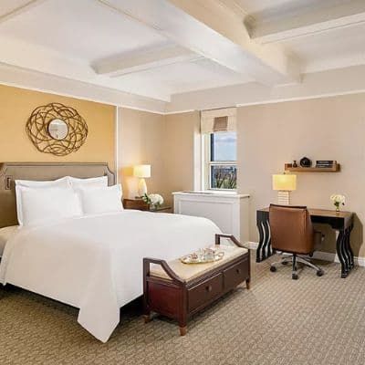 A hotel room at the Fairmont Le Château Frontenac featuring a large bed with white linens, a wooden bench, and a desk with a chair. The room has a warm, inviting color scheme.