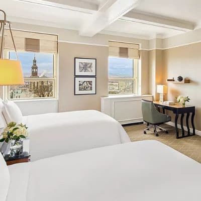 A hotel room at the Fairmont Le Château Frontenac featuring a large bed with white linens, a wooden bench, and a desk with a chair. The room has a warm, inviting color scheme.