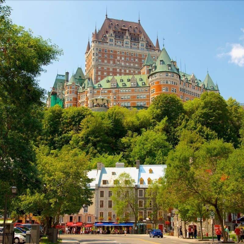 The historic Fairmont Le ChΓ’teau Frontenac hotel, with its red brick and green roofs, towers over a charming street in Quebec City, surrounded by lush green trees.