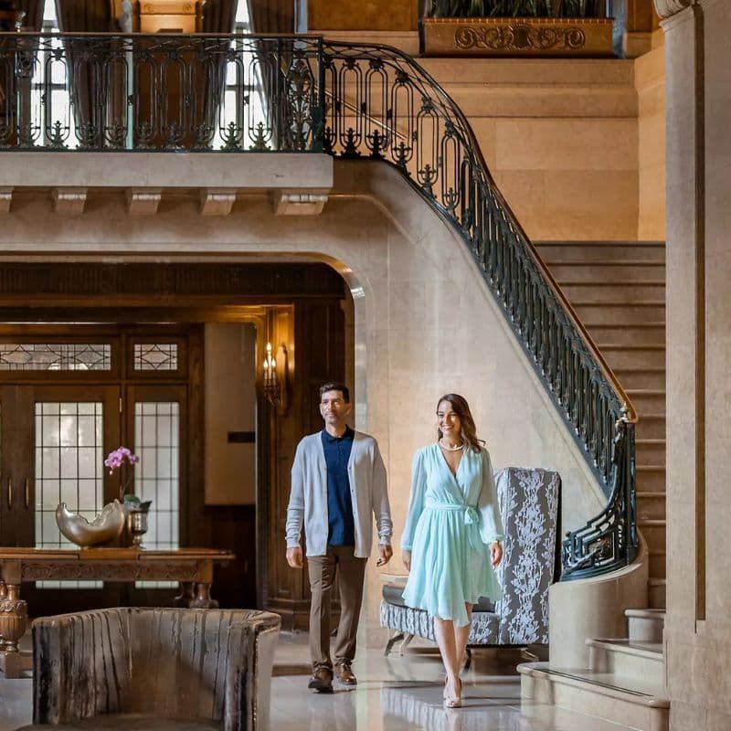 A couple walking through the grand, elegant lobby of a hotel, with a sweeping staircase and classic architectural details in the background.