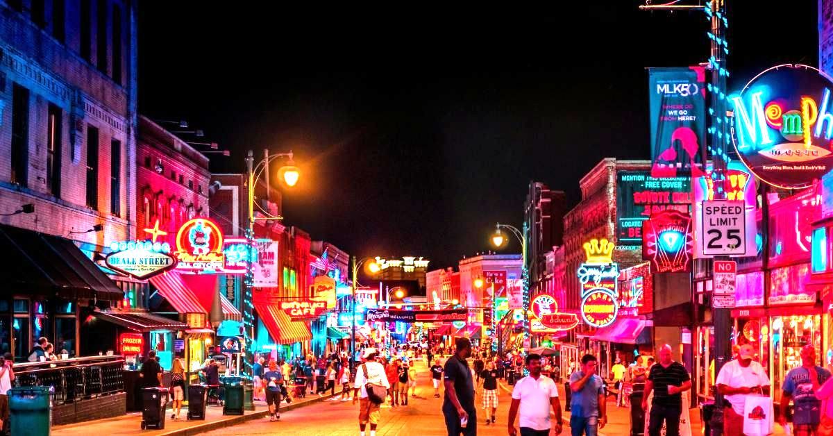 Group of senior travelers on a guided Memphis tour posing on Beale Street, with neon music venue signs and city skyline in the background.
