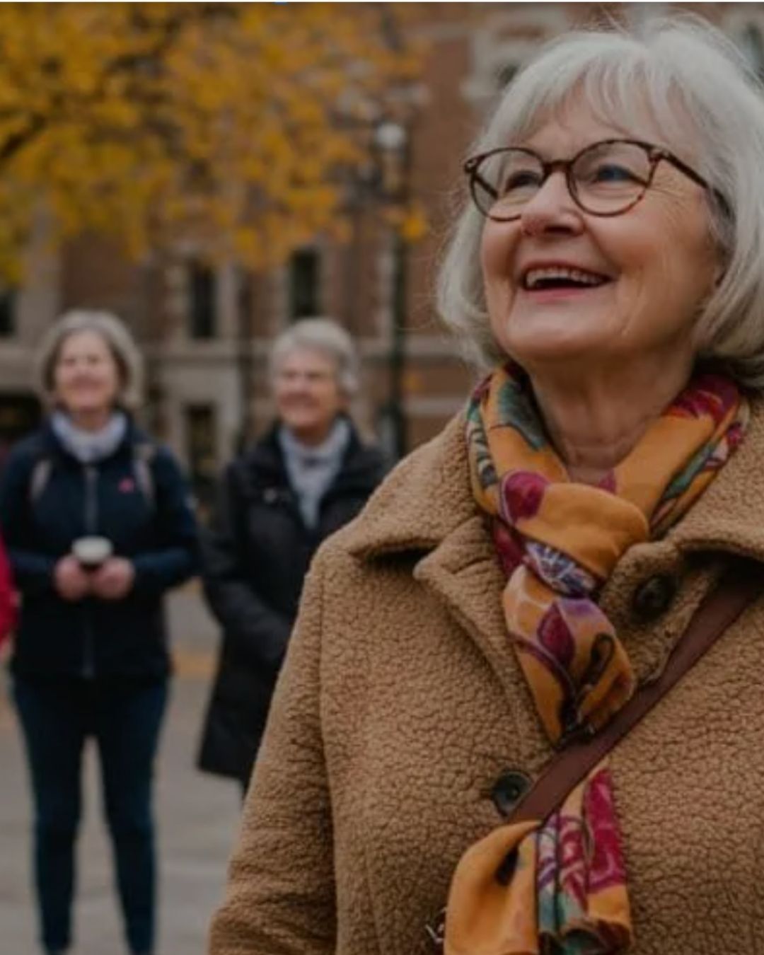 Smiling senior woman leading a small group of solo travelers during a guided walking tour in a historic city setting with autumn trees in the background.
