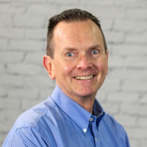 A man in a blue shirt is smiling in front of a white brick wall