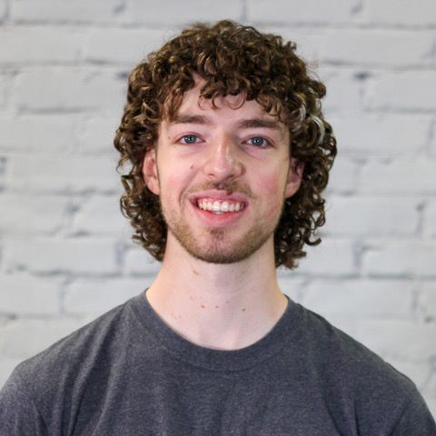 A young man with curly hair is smiling in front of a white brick wall.