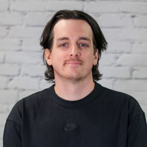 A man in a plaid shirt is smiling in front of a white brick wall