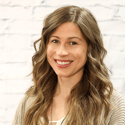 A woman with long hair is smiling in front of a white brick wall.