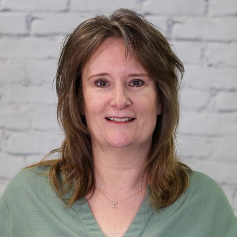 A woman in a blue shirt is smiling in front of a white brick wall.